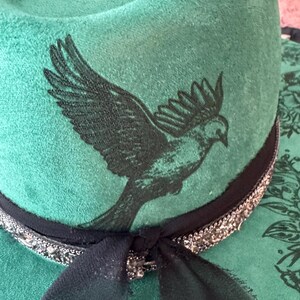 Puede incluir: Un sombrero de fieltro verde con una cinta negra y un dise&ntilde;o de p&aacute;jaro negro. El sombrero tiene una cinta negra con adornos plateados.