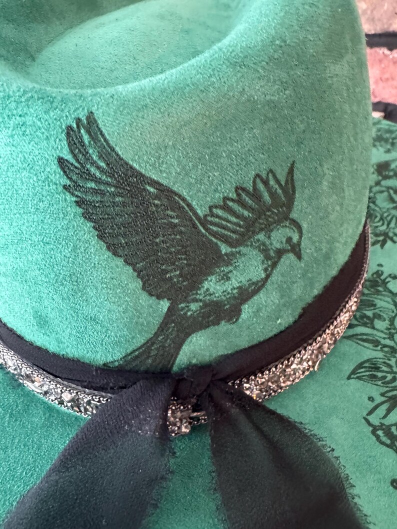 Puede incluir: Un sombrero de fieltro verde con una cinta negra y un dise&ntilde;o de p&aacute;jaro negro. El sombrero tiene una cinta negra con adornos plateados.