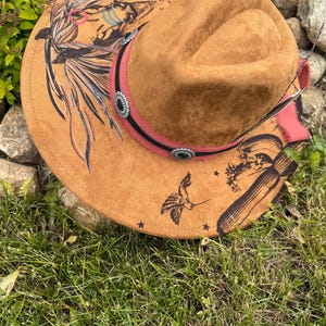 May include: A brown suede cowboy hat with a decorative band and silver-toned conchos. The hat features hand-painted designs including a hummingbird, feathers, and Native American-inspired artwork. The brim is adorned with small black stars.