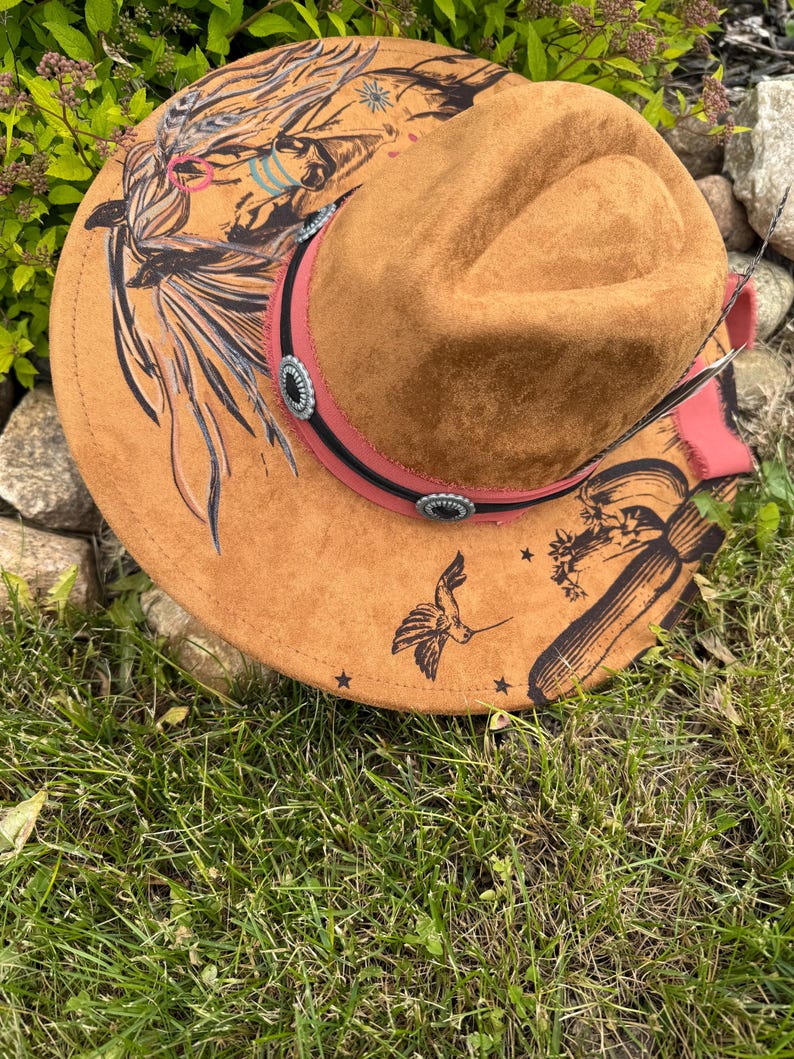 May include: A brown suede cowboy hat with a decorative band and silver-toned conchos. The hat features hand-painted designs including a hummingbird, feathers, and Native American-inspired artwork. The brim is adorned with small black stars.