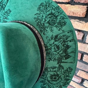 Puede incluir: Un sombrero fedora de fieltro verde con una banda negra y un ribete plateado. El sombrero est&aacute; decorado con dise&ntilde;os florales negros y el texto "It was".
