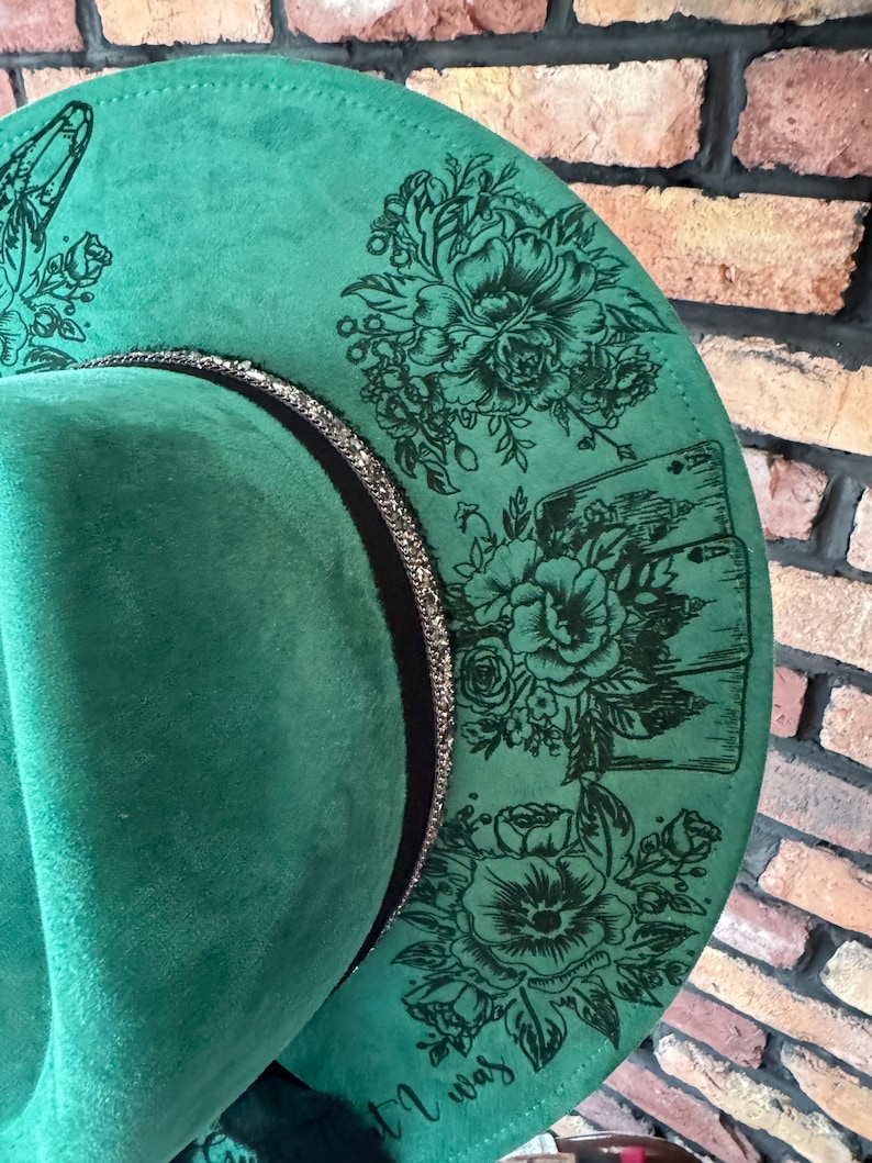 Puede incluir: Un sombrero fedora de fieltro verde con una banda negra y un ribete plateado. El sombrero est&aacute; decorado con dise&ntilde;os florales negros y el texto "It was".