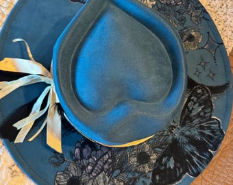 May include: A blue fedora hat with a wide brim, adorned with a butterfly and floral design. The hat features a ribbon and bow detail. The brim is decorated with a butterfly and floral pattern.