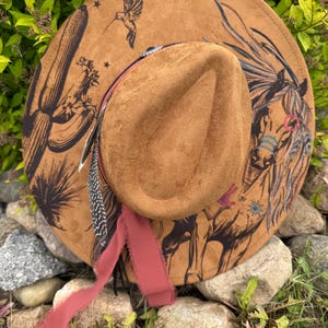 May include: A brown suede cowboy hat with a wide brim. The hat features a painted horse, cactus, and hummingbird design. A red ribbon and feathers are attached to the hat, adding a decorative touch.