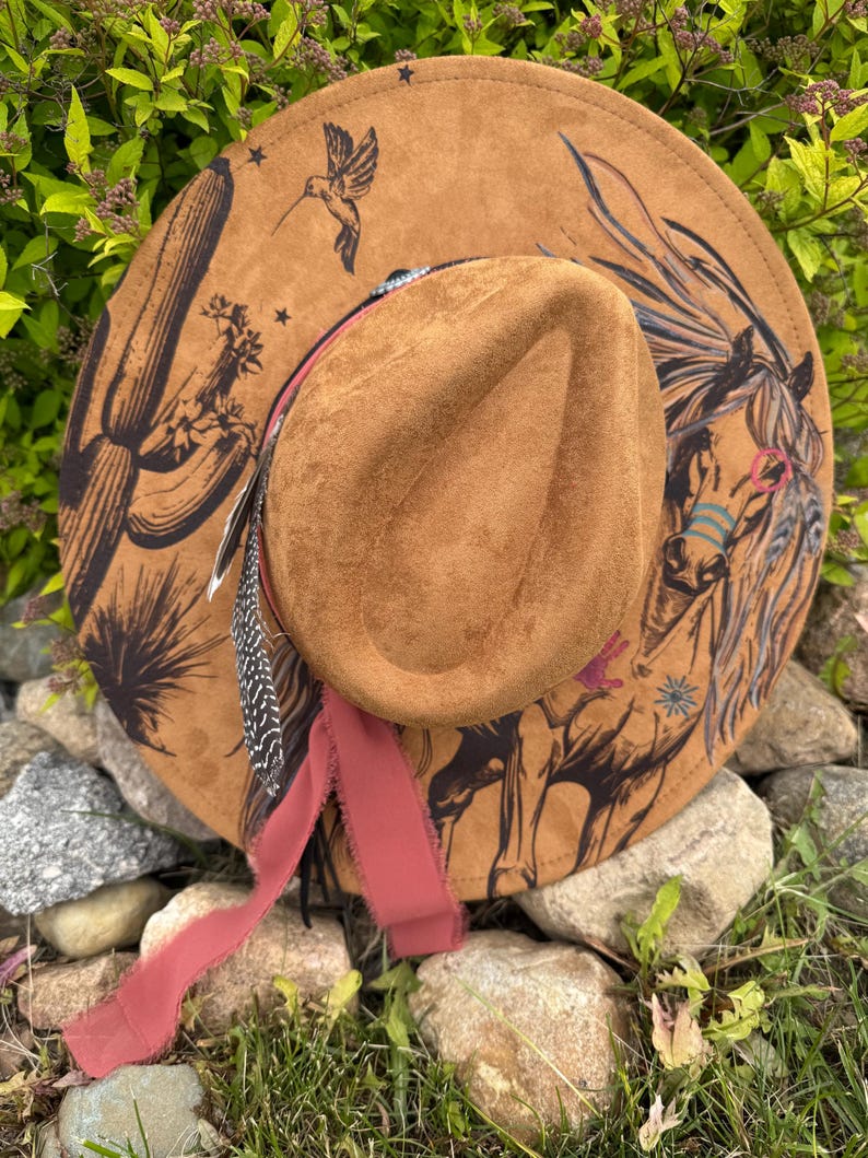 May include: A brown suede cowboy hat with a wide brim. The hat features a painted horse, cactus, and hummingbird design. A red ribbon and feathers are attached to the hat, adding a decorative touch.