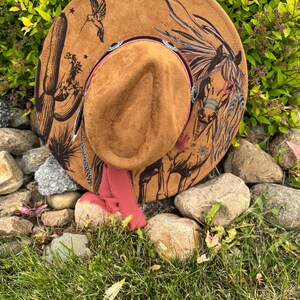 May include: A brown suede cowboy hat with a painted horse and desert scene. The hat features a decorative band with rhinestones and a pink ribbon. A hummingbird and cactus are also painted on the hat.