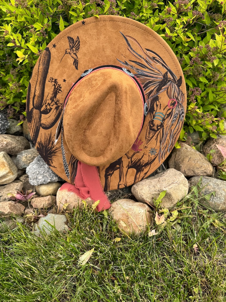 May include: A brown suede cowboy hat with a painted horse and desert scene. The hat features a decorative band with rhinestones and a pink ribbon. A hummingbird and cactus are also painted on the hat.