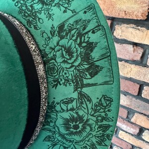 Puede incluir: Un sombrero de fieltro verde con una banda negra y un ribete de purpurina plateada. El sombrero est&aacute; decorado con un dise&ntilde;o floral y cartas de juego.