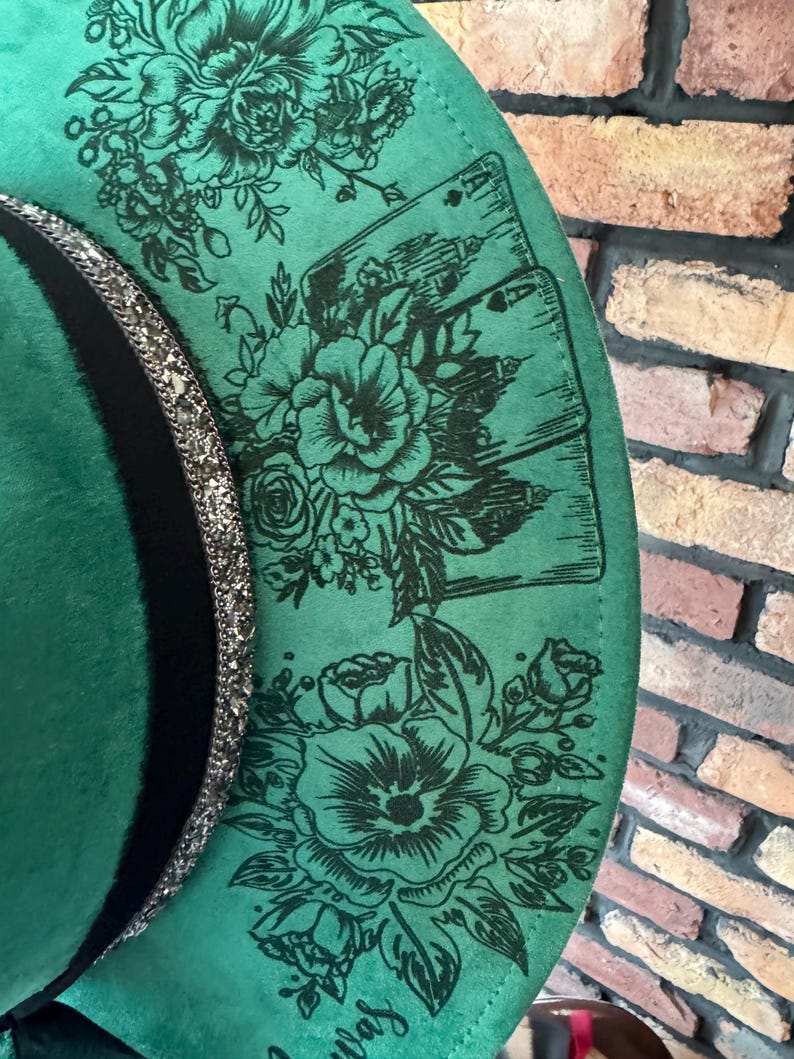 Puede incluir: Un sombrero de fieltro verde con una banda negra y un ribete de purpurina plateada. El sombrero est&aacute; decorado con un dise&ntilde;o floral y cartas de juego.