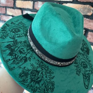 Puede incluir: Un sombrero fedora de fieltro verde con una banda negra y un ribete de purpurina plateada. El sombrero tiene un dise&ntilde;o floral con cartas de juego impresas en el ala.