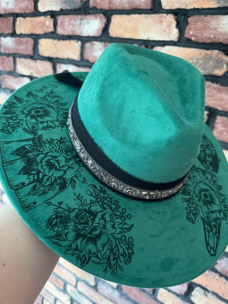 Puede incluir: Un sombrero fedora de fieltro verde con una banda negra y un ribete de purpurina plateada. El sombrero tiene un dise&ntilde;o floral con cartas de juego impresas en el ala.