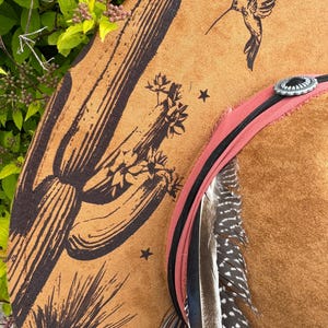 May include: A brown cowboy hat with a desert-themed design. The hat features a large cactus and hummingbird illustration, along with star accents. A pink and black band with feathers and a silver concho complete the look.
