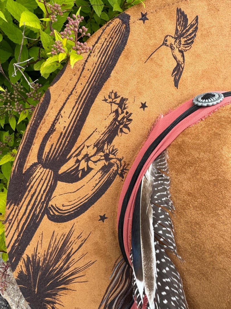 May include: A brown cowboy hat with a desert-themed design. The hat features a large cactus and hummingbird illustration, along with star accents. A pink and black band with feathers and a silver concho complete the look.