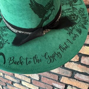 Puede incluir: Un sombrero fedora de fieltro verde con una cinta negra y un dise&ntilde;o de p&aacute;jaro. El sombrero tiene el texto "Back to the Gypsy that I was" escrito en el ala.