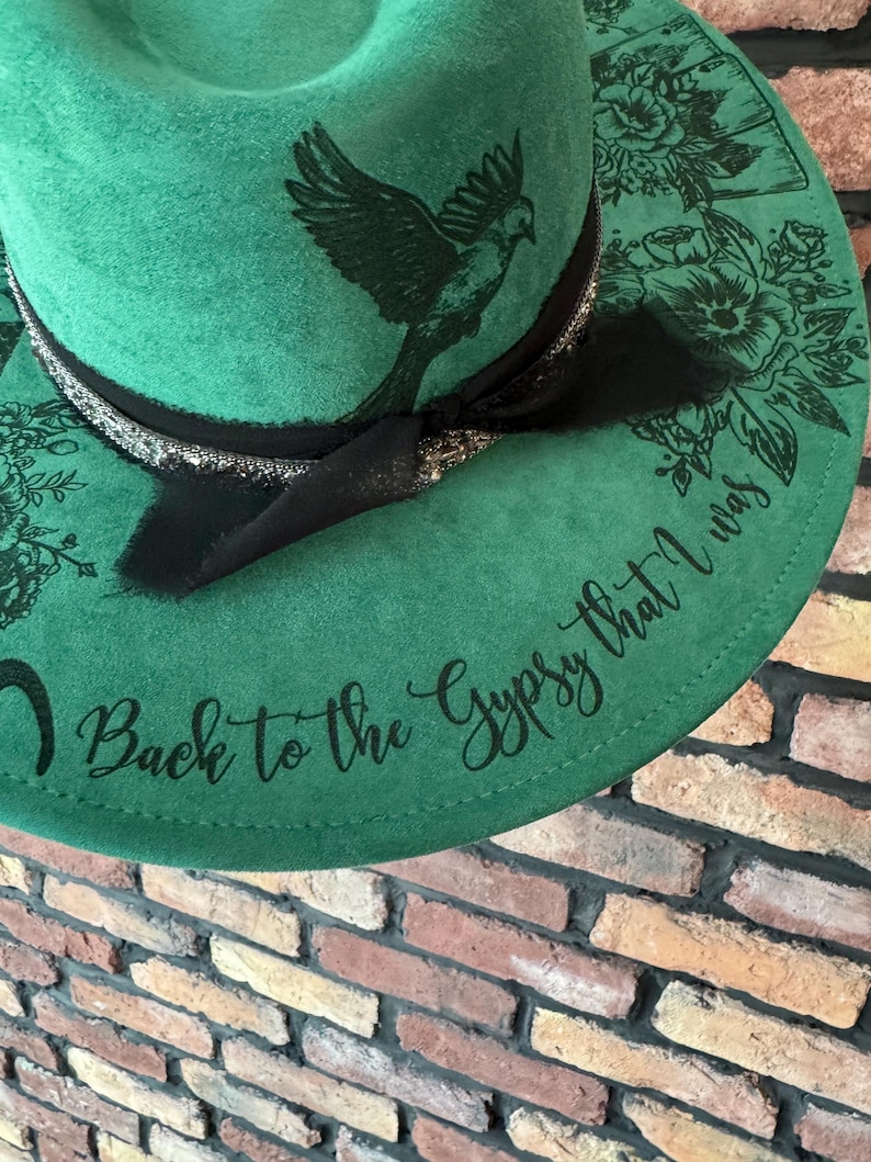 Puede incluir: Un sombrero fedora de fieltro verde con una cinta negra y un dise&ntilde;o de p&aacute;jaro. El sombrero tiene el texto "Back to the Gypsy that I was" escrito en el ala.