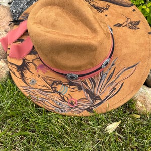 May include: A brown suede cowboy hat with a wide brim, featuring a hummingbird and floral design in black. The hat has a pink ribbon and decorative silver-toned accents around the crown.