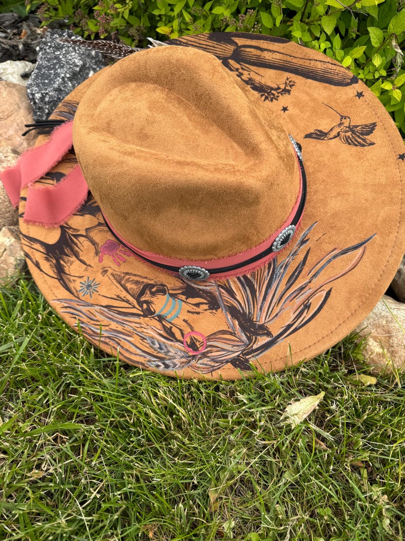 May include: A brown suede cowboy hat with a wide brim, featuring a hummingbird and floral design in black. The hat has a pink ribbon and decorative silver-toned accents around the crown.
