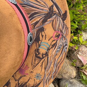 May include: A brown cowboy hat with a detailed horse illustration. The hat features a pink band with silver accents and a black stripe. The horse design includes colorful accents and hand-painted details, creating a unique, artistic look.