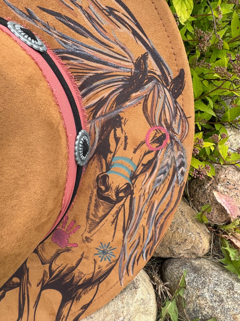May include: A brown cowboy hat with a detailed horse illustration. The hat features a pink band with silver accents and a black stripe. The horse design includes colorful accents and hand-painted details, creating a unique, artistic look.