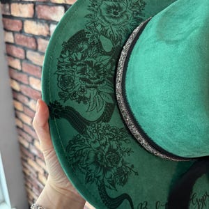 Puede incluir: Sombrero fedora de fieltro verde con un dise&ntilde;o de serpiente negra y flores. El sombrero tiene una cinta negra con purpurina plateada y las palabras "Back" y "Gypsy" escritas en el ala.