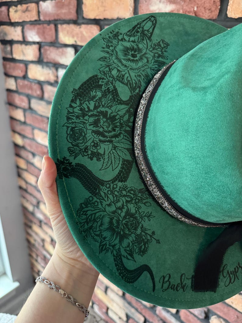 Puede incluir: Sombrero fedora de fieltro verde con un dise&ntilde;o de serpiente negra y flores. El sombrero tiene una cinta negra con purpurina plateada y las palabras "Back" y "Gypsy" escritas en el ala.