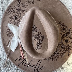 以下が含まれることがあります： 黒い花の模様と筆記体で「Michelle」という名前が入った、茶色のフェドラハット。 白い布のアクセントと羽、そして「2 Timothy 1:7」の文字が特徴です。