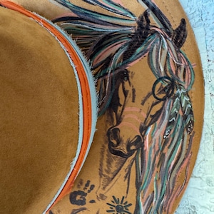 May include: A tan cowboy hat with a detailed illustration of a horse's head and colorful mane. The hat's brim features a handprint and sun design. The hat's interior trim is orange and light blue.
