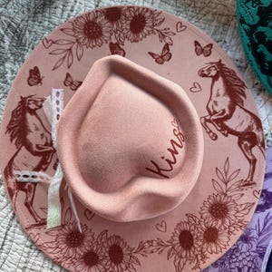 May include: A light pink hat with a heart-shaped crown featuring the name Kins embroidered in dark pink script. The wide brim is decorated with dark pink line art illustrations of horses, sunflowers, butterflies, and small hearts.