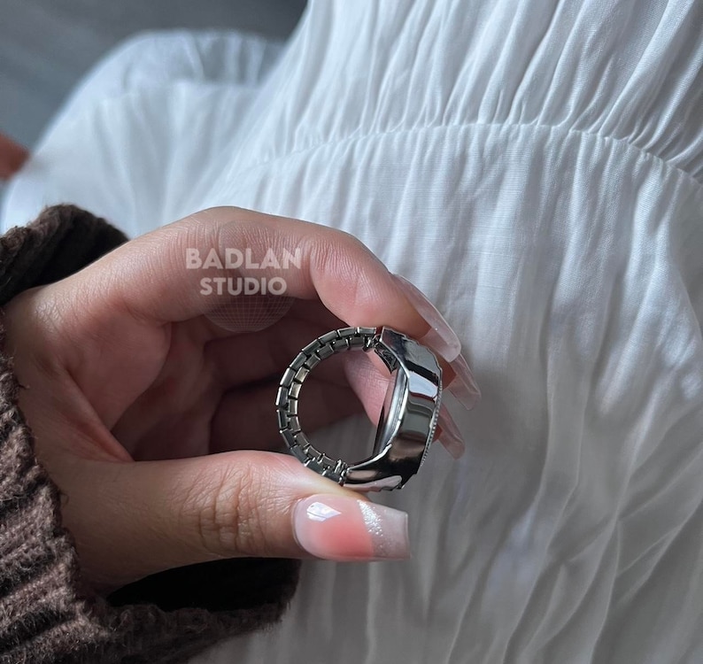 Puede incluir: Un anillo de reloj plateado con una esfera redonda y una banda plateada. La banda est&aacute; hecha de peque&ntilde;as piezas enlazadas que se asemejan a una cadena.