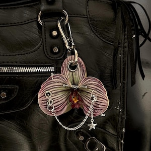 May include: A bag charm shaped like a striped purple and green orchid flower, featuring a silver-toned metal barbell piercing through the petals, a silver chain with a small star charm, and a metal lobster clasp for attachment to a handbag.