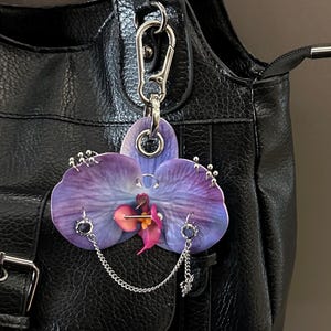 May include: A purple and blue orchid flower keychain featuring silver-toned metal piercings, including small rings, studs, and a delicate chain link. The floral ornament is attached to a silver metal lobster clasp hook for easy attachment.
