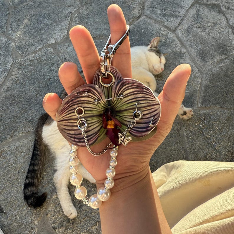 May include: A keychain featuring a flat, orchid-shaped acrylic charm with purple and green striped patterns. The charm is adorned with silver-toned metal rings, a small butterfly charm, a delicate silver chain, and a strand of white faux pearls.