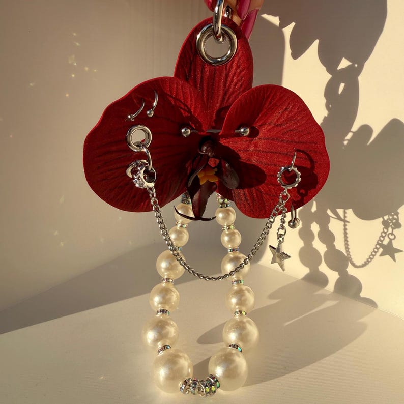 May include: A deep red artificial orchid flower adorned with silver-toned metal piercings, including hoops and a barbell. A silver chain connects the piercings, and a strand of white faux pearls with small silver star charms hangs from the flower.