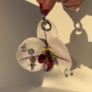 May include: A white orchid flower with purple speckles and a dark maroon center, featuring several silver-toned metal body jewelry pieces including rings, a star-shaped charm, and curved barbells attached directly to the flower petals.