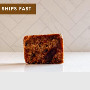 May include: A close-up of a dark brown bar of soap. The soap has a rough, textured surface and is rectangular in shape.