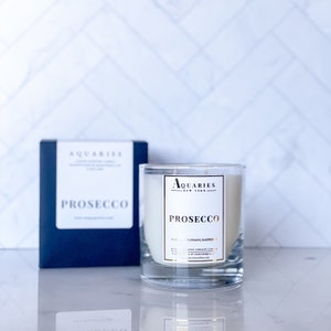 May include: A clear glass candle jar with a white candle inside. The candle is labeled "PROSECCO" with the brand name "AQUARIES NEW YORK" in gold lettering. The candle is sitting on a white surface with a blue box labeled "PROSECCO" in the background.