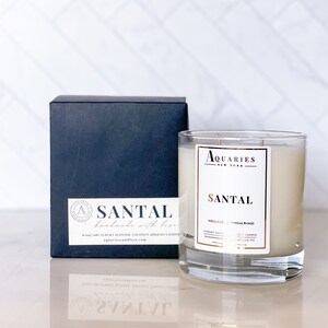Puede incluir: Un tarro de vidrio para velas con una vela blanca en su interior. El tarro tiene una etiqueta dorada que dice "Aquaries New York" y "Santal" con las notas de aroma "Shea + Sandalwood". La vela está hecha a mano con cera de coco y albaricoque y es una vela perfumada de lujo.