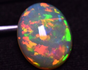 May include: Oval-shaped opal gemstone with a milky white base, displaying vibrant flashes of color. The opal's surface showcases a play of color, including red, orange, yellow, and green hues. The gemstone is set against a dark background.
