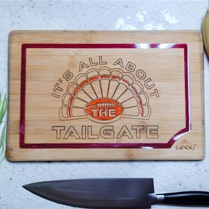 May include: A wooden cutting board with a red and white design. It features a turkey with a football inside and the text "It's All About The Tailgate". The board is made by Ginsu.