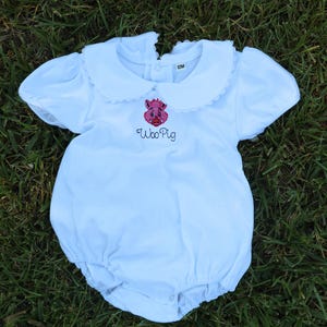 Puede incluir: Romper blanco para bebé con cuello Peter Pan y mangas cortas abullonadas. El romper presenta un cerdo rosa bordado y las palabras "Woo Pig" en escritura negra. La prenda está diseñada para un bebé de 12 meses.