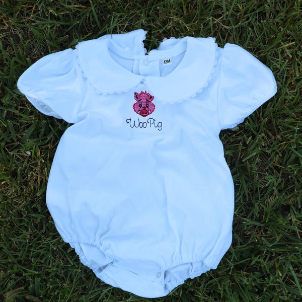 Könnte beinhalten: Weißer Baby-Strampler mit einem Bubikragen und kurzen, gepufften Ärmeln. Der Strampler zeigt ein gesticktes rosa Schwein und die Worte "Woo Pig" in schwarzer Schrift. Das Kleidungsstück ist für ein 12 Monate altes Baby konzipiert.