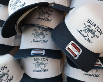 May include: A stack of beige and black trucker hats featuring a black embroidered logo that reads Burton Est. 2025 Social Club with an illustration of two mugs. Each hat has a black brim and a rectangular Otto brand sticker on the front.