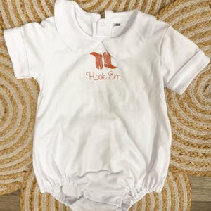May include: White baby romper with a Peter Pan collar and short sleeves. The romper features an embroidered design of orange cowboy boots and the words "Hook Em". The size is 18M.