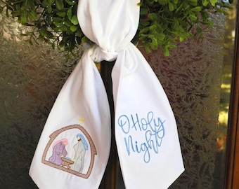 Könnte beinhalten: Eine weiße Stoff-Hängedekoration mit einer gestickten Krippenszene auf der einen Seite und dem blau gestickten Text O Holy Night auf der anderen. Der Stoff ist oben verknotet und zeigt einen braunen Stall, einen gelben Stern und Figuren.