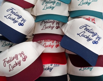 May include: A collection of baseball caps with the phrase "Feeling Lucky" embroidered on the front. The caps have a white crown and a colored brim, with options including red, blue, and green. Some caps feature a cherry graphic.
