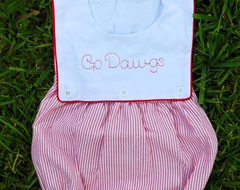May include: A white baby romper with red trim and the embroidered text "Go Dawgs". The romper features a red and white striped bottom and three white buttons. The garment is set against a backdrop of green grass.