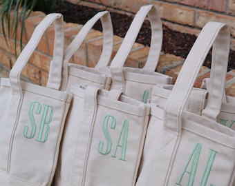 May include: A group of off-white canvas tote bags with two sturdy handles. Each bag features a unique two-letter monogram embroidered in light green thread on the front. The visible monograms include the letters SB, SA, and AL in a vertical font.