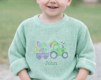 May include: A young child wearing a light green knit sweater with a tractor and trailer design embroidered on the front. The tractor is green, and the trailer is purple. The name "John" is embroidered below the design. The child is wearing blue jeans.