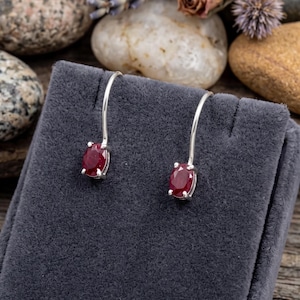May include: Silver drop earrings featuring oval-cut, deep red gemstones. The earrings are presented on a grey velvet jewellery stand, with a backdrop of stones, dried flowers, and lavender sprigs.