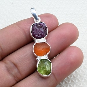 Puede incluir: Colgante de plata con tres piedras preciosas de colores: un púrpura oscuro, un naranja y un verde claro. Las piedras tienen forma irregular y están engastadas en biseles individuales de plata. El colgante tiene un lazo de plata en la parte superior.